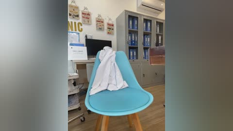 Video of a modern, brightly-lit medical office with a turquoise Eames-style chair, white coat draped over it, against a white wall with framed medical posters, grey cabinets, and a white AC unit.