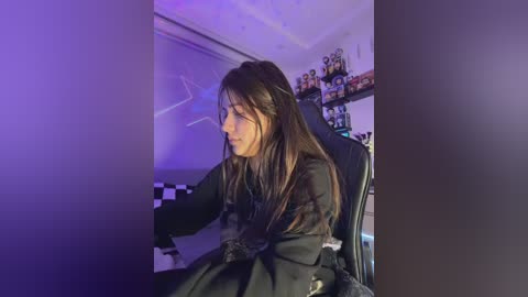 Video of a young woman with long, straight brown hair, sitting in a black gaming chair, playing a video game in a dimly lit room with purple lighting, checkered floor, and shelves filled with gaming figurines in the background.