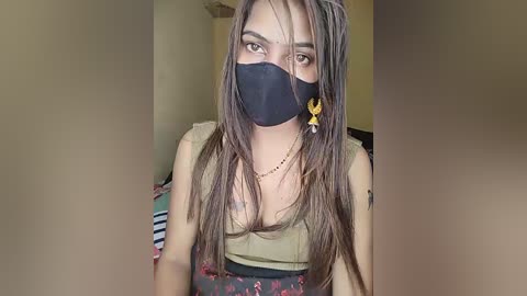 Video of a young woman with long, straight brown hair, wearing a black face mask, olive-green tank top, and a floral-patterned skirt. She has a light skin tone, and a yellow earring dangles from her left ear. Background shows a beige wall and a striped blanket.