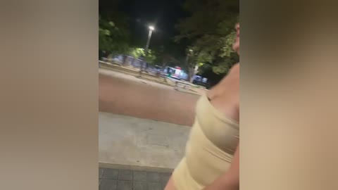 Video of a blurred woman in a beige tank top walking on a sidewalk at night. Background shows a street with parked cars, streetlights, and trees.