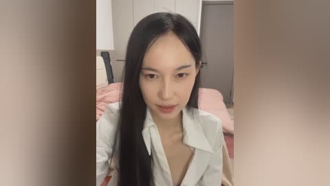 Video of a young Asian woman with long black hair, wearing a light blue shirt, sitting on a bed with pink sheets, in a softly lit bedroom.