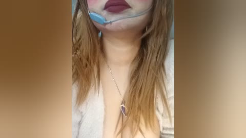 A close-up video of a woman with fair skin, long wet hair, wearing a blue ball gag, maroon lipstick, and a silver necklace with a purple pendant, blurred background.