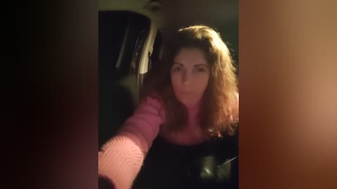 A video of a woman with long, curly brown hair, wearing a pink sweater, smiling while holding a black purse. The background is dimly lit, with a blurred, dark figure partially visible.