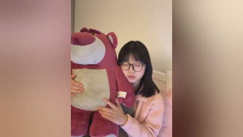 A video of a woman with glasses, light skin, and shoulder-length black hair, wearing a pink sweater, hugging a plush, red, cartoon bear with a white belly. The background is a plain, off-white wall.