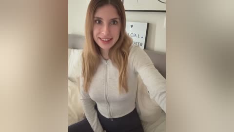 Video of a smiling, fair-skinned woman with long, straight blonde hair, wearing a white zip-up top and black leggings, sitting on a bed with beige bedding.