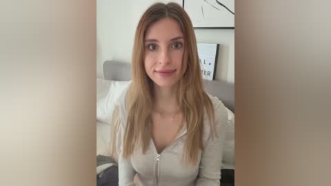 A video of a young Caucasian woman with light skin, straight blonde hair, and a slender physique, wearing a light gray zip-up hoodie. She is indoors, sitting on a bed with white sheets, and a white wall with a framed black and white abstract art piece in the background.