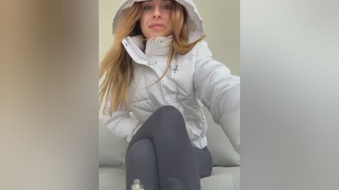 Video of a young woman with long, straight, light brown hair, wearing a white puffy jacket, gray leggings, and sitting on a white leather chair. The background is plain and neutral.
