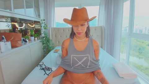 Video of a slender, tattooed woman with light skin in a modern, sunlit room, wearing a tan cowboy hat and gray crop top.