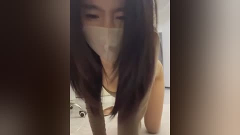 A video of a young Asian woman with long black hair, wearing a white mask, and a gray tank top, crouching on a gray carpet. The background includes a white wall and a metallic object.