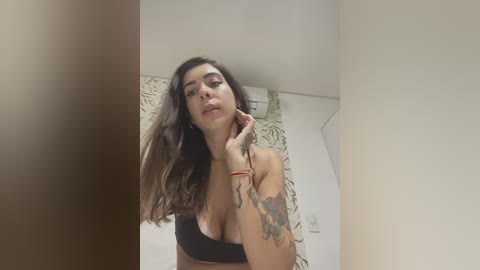 A video of a tattooed woman with long brown hair, wearing a black bra, standing in a bathroom with a floral shower curtain.