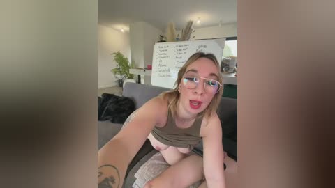 A video of a fair-skinned, slender woman with glasses, wearing glasses, in a beige tank top, sitting on a grey couch, with a whiteboard in the background, and a blurred, naked male figure in the foreground.