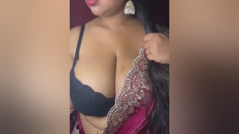 Video of a South Asian woman with medium-brown skin and long black hair, wearing a black lace bra and a red embroidered sari. She is seen from the shoulders down, with her hand holding the sari.