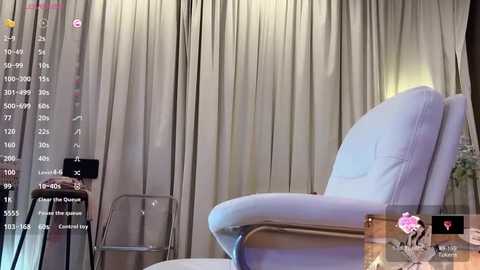 Video of a modern living room with beige curtains, a white leather armchair, and a minimalist metal chair. A video feed displays an Asian woman in a pink dress sitting on the armchair.