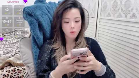 A video of an Asian woman with long, dark hair and light skin, wearing a black sweater, sitting on a bed with a leopard-print blanket, taking a selfie with a pink phone.