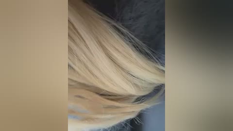 A close-up video of blonde hair, featuring soft, wavy strands with natural highlights. The background is blurred, with shades of grey and beige, focusing attention on the hair texture and color.