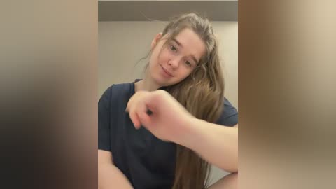 Video of a young Caucasian woman with long brown hair, wearing a navy blue t-shirt, making a fist in a playful manner. Background is blurred and neutral-toned.