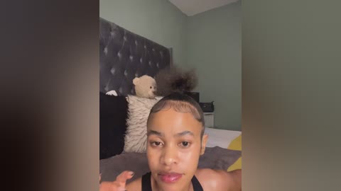 Video of a young African American girl with a short Afro, lying on a bed in a dimly lit room with green walls. She has a neutral expression and is wearing a black tank top.
