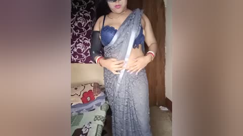 Media: Video of a South Asian woman in a blue, lace-trimmed sari and matching bra, standing in a room with patterned curtains and a bedspread.