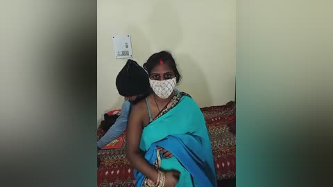 Video of an Indian woman with medium skin tone, dark hair, and a blue saree, wearing a face mask and red bindi, sitting on a patterned bedspread against a plain beige wall.