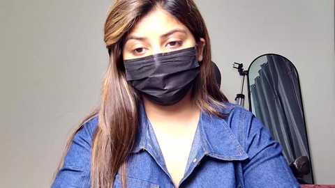 Video of a South Asian woman with long, straight, light brown hair wearing a black face mask, denim shirt, and gray walls.