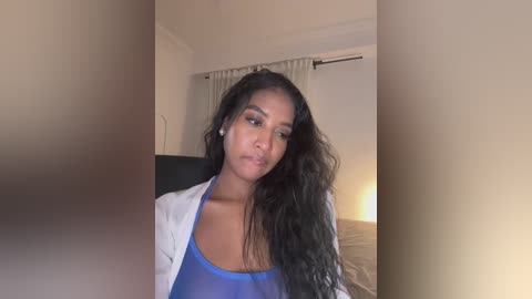 Video of a young Black woman with long, wavy black hair, wearing a blue tank top and white cardigan, sitting indoors in a dimly lit room with cream-colored walls and a closed window.