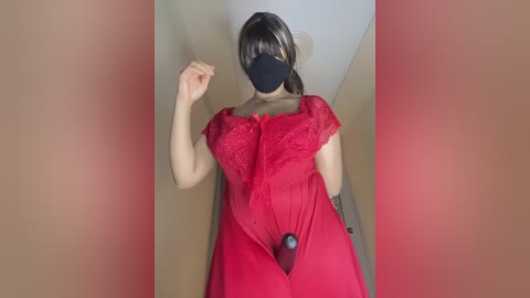 Video of a woman in a red lace dress, black mask, and a black dildo attached to her vagina, taken indoors with blurred background.