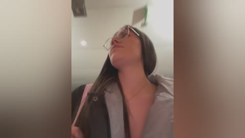 Video of a woman with long brown hair, wearing glasses, a pink shirt, and gray cardigan, leaning against a white wall in a dimly lit corridor.