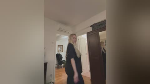 Video of a blonde woman in a black dress standing in a dimly lit, sparse apartment with white walls, wooden floors, and open doorways.