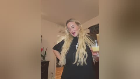 Video of a blonde woman in a black shirt, holding a glass, in a warmly lit, sparsely decorated room with beige walls and wooden floors.