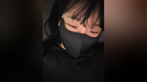 A video of a person with short black hair, wearing a black mask, in a dimly lit room, eyes obscured, creating a sense of mystery and anonymity.