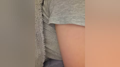 A video showcasing a close-up of a person's bare arm and hand, with a grey short-sleeved shirt and a fluffy, grey blanket partially covering the shoulder. The background is neutral and blurred, focusing on the texture of the fabric and the softness of the blanket.
