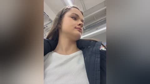 Video of a young woman with light skin and straight brown hair, wearing a white ribbed sweater and black jacket, standing in an airplane. The background shows a blurry cabin interior with overhead lights and air vents.