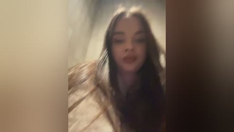 Video of a woman with long, wavy brown hair, wearing a dark top, standing in a dimly lit room. The background is blurred, emphasizing her face and hair.