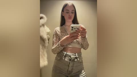 Video of a young woman with light skin, straight brown hair, wearing a leopard print crop top and high-waisted jeans, taking a selfie in a beige room, holding a white iPhone.