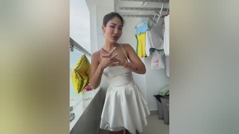 Video of a young Asian woman with medium build, fair skin, and dark hair in a white sleeveless dress, holding a yellow garment, standing in a modern, bright laundry room with hanging clothes.