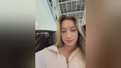 Video of a young Caucasian woman with light skin, long blonde hair, and closed eyes, seated in a modern, spacious interior with high ceilings and glass walls, wearing a light-colored hoodie.