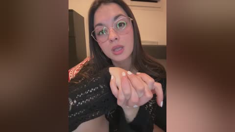 A video of a young woman with long, dark hair, wearing glasses, a black lace top, and a red-patterned cardigan, making a heart shape with her hands in a cozy indoor setting.