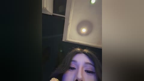 A video of a woman with long black hair, seen from above, capturing her face and upper body. The background is dimly lit with a white ceiling light, creating a moody, intimate atmosphere.