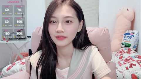 Video of a young Asian woman with long black hair, wearing a light pink top, sitting on a bed with a pink teddy bear and heart-patterned blanket.