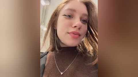 Video of a young Caucasian woman with fair skin, shoulder-length auburn hair, wearing a brown ribbed turtleneck sweater and dangling silver earrings, indoors with blurred background.