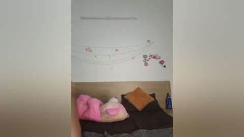 Video of a minimalist room with a white wall featuring a decorative string of pink and black circular stickers. A beige sofa with a brown cushion and a pink stuffed toy is in the foreground.