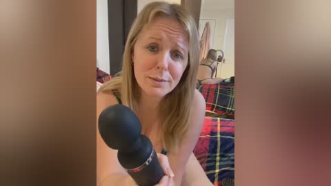 Video of a blonde woman with a microphone, recording a podcast in a cozy, plaid-patterned bedroom.