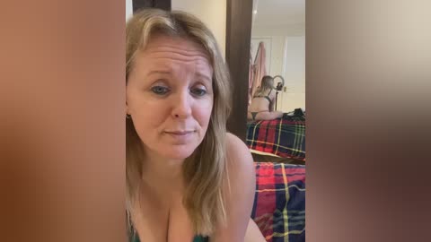 A candid video captures a blonde woman with fair skin and blue eyes, wearing a green top, looking through a mirror. Behind her, a plaid bedspread and a cluttered room are visible.
