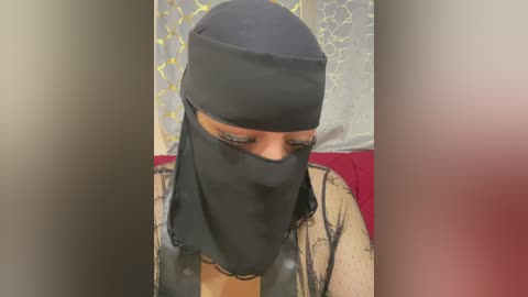 Video of a woman with light brown skin wearing a black niqab, sitting indoors against a gray and yellow-patterned background, partially visible. She has a beige, lace-trimmed top.