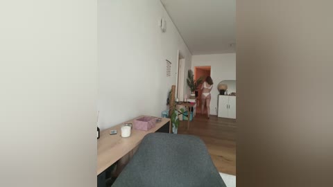 A video of a modern, minimalist living room with white walls, light wooden floors, and a small wooden table holding a vase and a cup. A person in pink underwear stands in the doorway to the right.