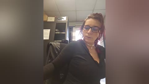 Video of a woman with fair skin and red hair in glasses, wearing a black top and pearl necklace, seated in an office with bookshelves, computer monitor, and desk.