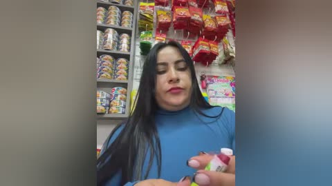 Video of a Latina woman with long black hair, wearing a blue turtleneck, holding a pack of condoms in a brightly lit convenience store. Shelves behind her are filled with colorful candy and snacks.