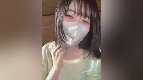 Video of an East Asian woman with straight black hair, wearing a white face mask, light yellow shirt, and closed eyes, in a wooden room.