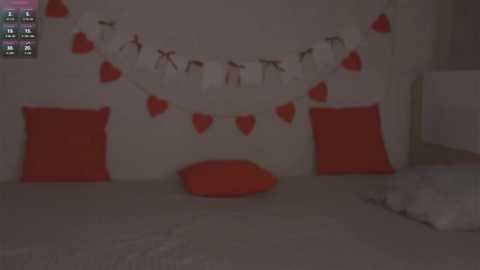 A dimly lit video of a bedroom with a white wall adorned with a heart-shaped banner and red heart-shaped cushions on a bed, featuring a soft, orange floor cushion.