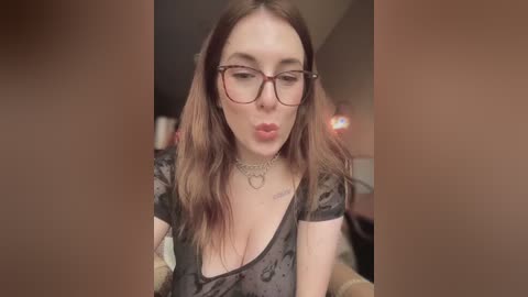 A video of a fair-skinned woman with long brown hair and glasses, wearing a black floral dress, blowing a kiss, with a blurred background.